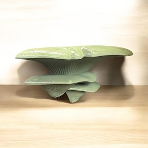 May include: A light green mushroom sculpture with a layered design. The sculpture has a textured surface, with each layer resembling a mushroom cap. The artwork is displayed on a light wooden surface.
