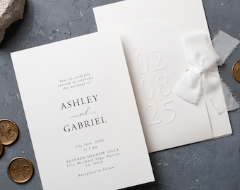 Puede incluir: Elegante juego de invitaciones de boda blancas con detalles en relieve y una delicada cinta. La invitación incluye los nombres Ashley y Gabriel, la fecha 16 de julio de 2025 y los detalles del lugar. Sellos de cera dorados y piedras decorativas completan el diseño.