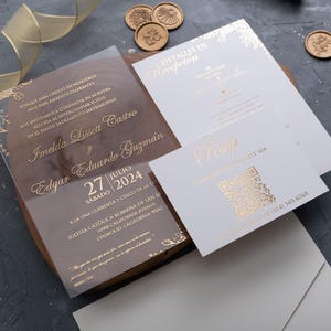 Op de afbeelding: Trouwuitnodigingsset met een heldere acryl uitnodiging met gouden letters, een witte receptiekaart en een RSVP-kaart met een QR-code. De set bevat een houten basis, gouden zegels en een gouden lint.