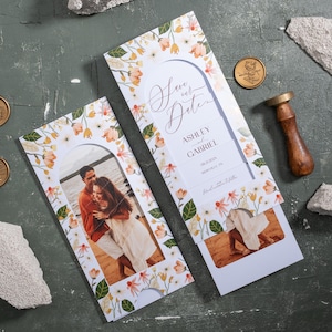 May include: Wedding "Save the Date" cards with floral designs and photos of a couple. The cards feature the names Ashley and Gabriel, along with the date and location. Gold wax seals and a wooden stamp are also present.