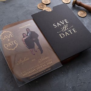 Op de afbeelding: Elegant trouwkaarten set met een heldere acrylkaart met gouden tekst en een foto van het paar, en een zwarte envelop. De woorden "Save the Date" staan prominent afgebeeld.