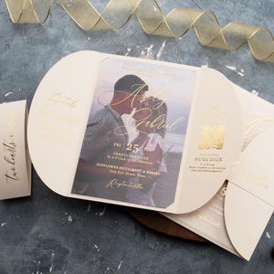 Puede incluir: Conjunto de invitaciones de boda en marfil con letras doradas. La invitación presenta una foto de una pareja besándose, con los nombres "Ashley & Gabriel" en una elegante escritura. Incluye detalles de la fecha, hora y lugar de la boda.