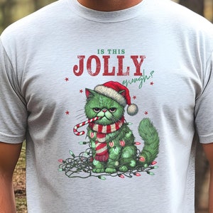 May include: Pale grey t-shirt with a grumpy green cat wearing a Santa hat, scarf, and Christmas lights. The cat holds a candy cane. Text on the shirt reads "IS THIS JOLLY enough?" A festive Christmas design.