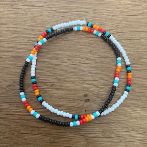 Op de afbeelding: Twee kralenkettingen, een zwarte, een witte met oranje, rode en turquoise kralen.