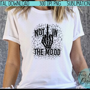 May include: A white t-shirt with a black skeleton hand giving the middle finger, with the words "Not in the mood" in black boxes on a leopard print background.