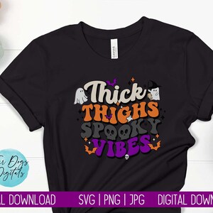 Op de afbeelding: Zwart T-shirt met een Halloween-thema grafische afbeelding met de tekst "Thick Thighs Spooky Vibes" in kleurrijke letters. De afbeelding bevat ook geesten, vleermuizen en schedels.