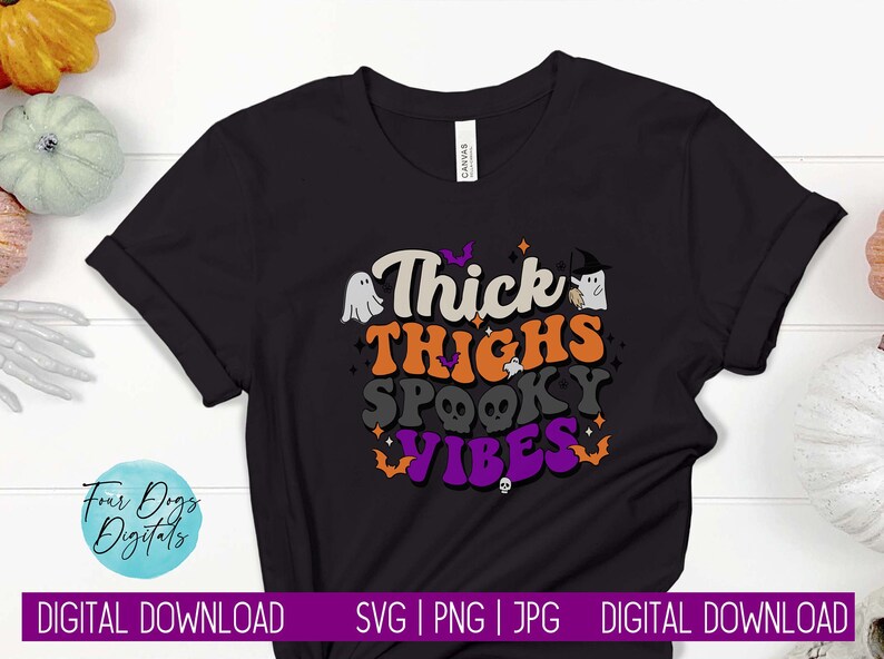Op de afbeelding: Zwart T-shirt met een Halloween-thema grafische afbeelding met de tekst "Thick Thighs Spooky Vibes" in kleurrijke letters. De afbeelding bevat ook geesten, vleermuizen en schedels.