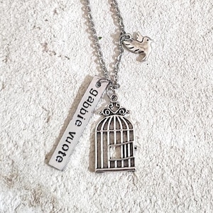 Può includere: Una collana argentata con un ciondolo a forma di gabbia per uccelli, un piccolo ciondolo a forma di uccello e un'etichetta con la scritta "gabbie vuote". La collana è presentata su una superficie strutturata bianco sporco.