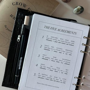 Op de afbeelding: Een zwarte map met een getextureerde kaft bevat een witte pagina met de titel THE FIVE AGREEMENTS. De pagina somt vijf genummerde principes met affirmaties op, waaronder Be Impeccable With Your Word en Don't Take Anything Personally. Een zwarte pen is bevestigd.