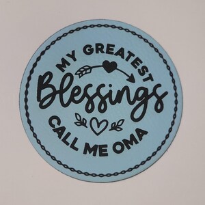 May include: A round, light blue decorative item with the text "MY GREATEST Blessings CALL ME OMA" in black script. The text is arranged around a heart and arrow design. The item has a black dotted border.