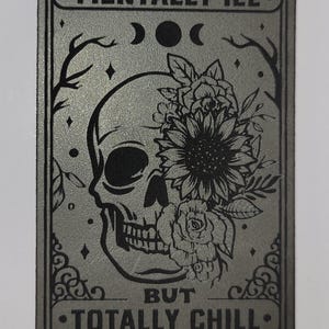 May include: A rectangular, gray metal sign with the words "MENTALLY ILL" and "BUT TOTALLY CHILL" in black. A skull with floral accents is in the center. The design includes moons, stars, and decorative flourishes.