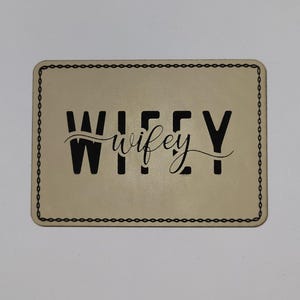 May include: A rectangular coaster with the word "WIFEY" in black, stylized text. The word "wifey" is in a cursive font. The coaster has a light beige background and a black chain-link border.
