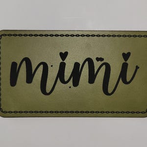 May include: Olive green rectangular patch with the word "mimi" in black cursive script. Two small black hearts are above the "i" letters. The patch has a black chain-link border.