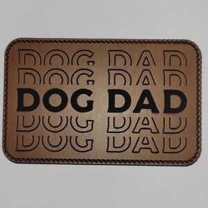 May include: A rectangular, brown leather patch with the words "DOG DAD" in bold, black letters. The phrase is repeated multiple times in a stacked pattern. The patch has a decorative rope-like border.