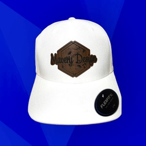 May include: White baseball cap with a brown leather patch featuring the text "Mavery Design" in black lettering. The patch has a hexagonal shape with a floral design. The cap has a black Flexfit label on the side.