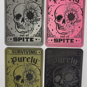 May include: Four rectangular cards with a skull design. Each card features the words "SURVIVING" and "Purely" above a skull with floral accents. The phrase "out of SPITE" is at the bottom. The cards are silver, pink, olive green, and black.