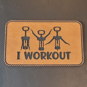 May include: A rectangular, tan leather patch with the phrase "I WORKOUT" in black, stylized text. The design features three black corkscrew illustrations. The patch has a dark brown rope-like border.