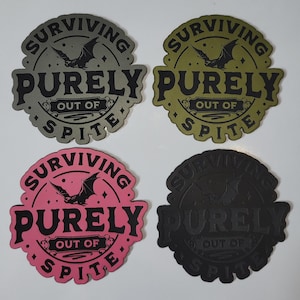May include: Four round stickers with the text "SURVIVING PURELY OUT OF SPITE" and a bat illustration. The stickers are in gray, olive green, pink, and black.
