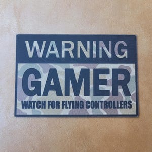 May include: A rectangular patch with the words "WARNING GAMER" in white letters over a camouflage pattern. The text "WATCH FOR FLYING CONTROLLERS" is below. The patch is set against a tan background.
