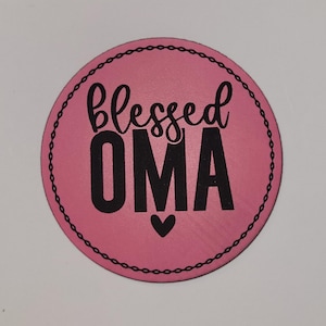 May include: A round, pink item with the words "blessed OMA" in black script and block letters, with a small heart below the word "OMA". The item has a black chain-like border.