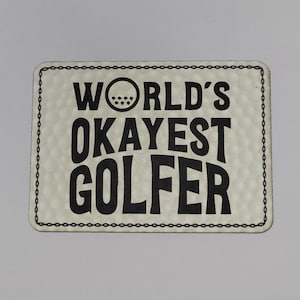 May include: A rectangular sign with the text "WORLD'S OKAYEST GOLFER" in black, bold letters. The sign has a textured, off-white background and a black chain-link border. A golf ball graphic is above the word "OKAYEST".