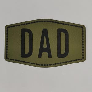 May include: Olive green patch with the word "DAD" in bold black letters. The patch has a stitched border and a unique, slightly curved shape. The design is simple and clear, making it easily readable.