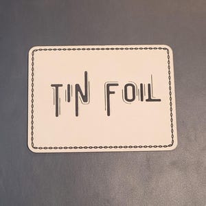 May include: A rectangular, off-white sticker with the words "TIN FOIL" in black, stylized font. The sticker has a black dotted border and is set against a gray background.