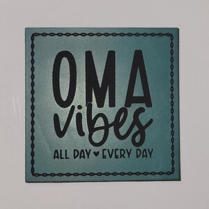 May include: Square teal sign with the words "OMA vibes" in large black letters, with "ALL DAY EVERY DAY" below. The sign has a black stitched border and is on a white background.