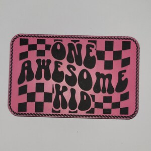 May include: A pink rectangular patch with a black and white checkerboard pattern and the words "ONE AWESOME KID" in black bubble letters. The patch has a decorative rope-like border.
