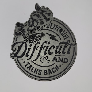 May include: A silver-colored, circular sticker with the words "Difficult & Talks Back" in a stylized font. A skeleton figure with a beer mug is depicted above the text, with the word "Expensive" around the top edge.