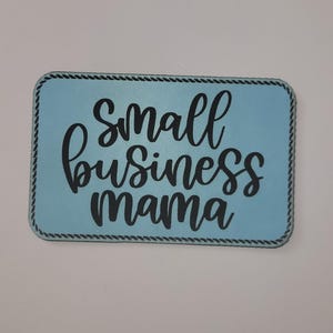 May include: A rectangular sign with a light blue background and black text that reads "Small Business Mama." The sign has a black rope-like border.