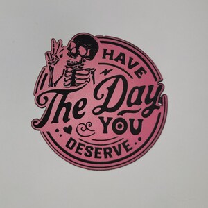 May include: A pink and black circular sticker with the text "Have The Day You Deserve." A skeleton is giving a peace sign. The sticker has a vintage, gothic aesthetic.