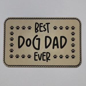 May include: A rectangular sign with rounded corners, featuring the text "BEST DOG DAD EVER" in bold black letters. The sign is beige with a black rope-like border and paw prints around the edges.