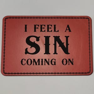 May include: A rectangular, red sign with the text "I FEEL A SIN COMING ON" in black, stylized lettering. The sign has a black chain-link border.