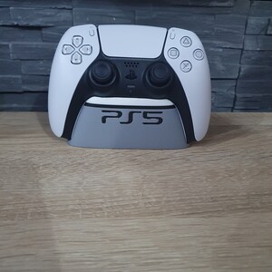 Op de afbeelding: Een witte PlayStation 5-controller staat op een grijze standaard met het woord "PS5" erop gedrukt.