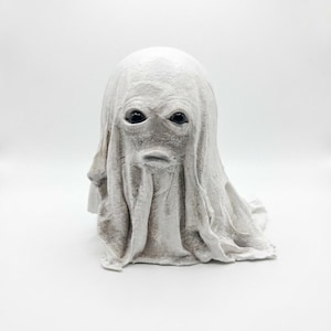 May include: A white, textured ghost figurine with a draped fabric appearance. The sculpture features two dark, glossy, round eyes and a small, horizontal mouth opening, with folds and creases mimicking a sheet covering a rounded form.