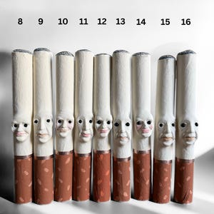 Puede incluir: Ocho esculturas de cigarrillos hechas a mano, cada una con una cara tallada única. Los cigarrillos tienen tubos de papel blanco, filtros marrones y puntas grises. Los números del 8 al 16 están impresos arriba. Una pieza de arte peculiar.