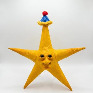May include: A yellow five-pointed star figure with a textured surface and a human-like face featuring dark eyes and a nose. The star wears a small, pointed blue crochet hat with a yellow ruffled brim and a red pom-pom on the very top point.