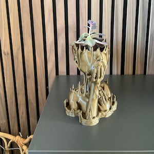 Könnte beinhalten: Ein goldfarbener, 3D-gedruckter Pflanztopf in Form eines menschlichen Kopfes mit einer kleinen Sukkulente, die aus der Oberseite wächst.