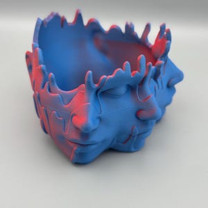 Könnte beinhalten: Ein blauer und roter 3D-gedruckter Pflanztopf in Form eines Gesichts mit einem geschmolzenen oder tropfenden Effekt.