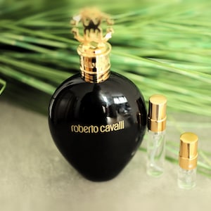 Puede incluir: Un frasco de perfume de vidrio negro con una tapa dorada y una etiqueta dorada que dice "roberto cavalli". El frasco está sobre una superficie gris junto a dos frascos de perfume más pequeños con tapas doradas.