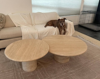 May include: Two beige travertine coffee tables with round tops and cylindrical bases. The tables are different sizes and sit on a patterned rug. A brown dog rests on a cream-colored sofa in the background.