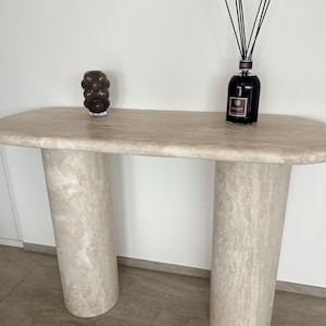 May include: A white travertine console table with two round legs. The table has a light brown, textured surface. A brown glass vase with a cluster of brown glass spheres sits on the table. A glass bottle with black reeds sits on the table.