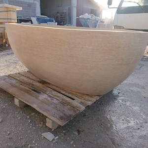 May include: A large, beige stone bathtub with a rounded, bowl-like shape. The tub sits on a wooden pallet, showcasing its size and texture. The background includes construction materials and a truck.