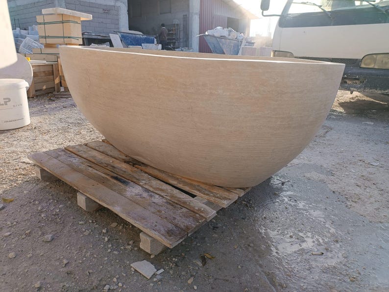 May include: A large, beige stone bathtub with a rounded, bowl-like shape. The tub sits on a wooden pallet, showcasing its size and texture. The background includes construction materials and a truck.