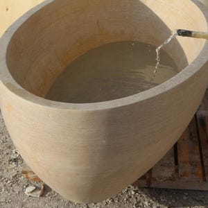 May include: A large, beige stone bathtub with an oval shape. Water flows into the tub from a hose. The tub sits on a wooden pallet, and the background is a construction site.
