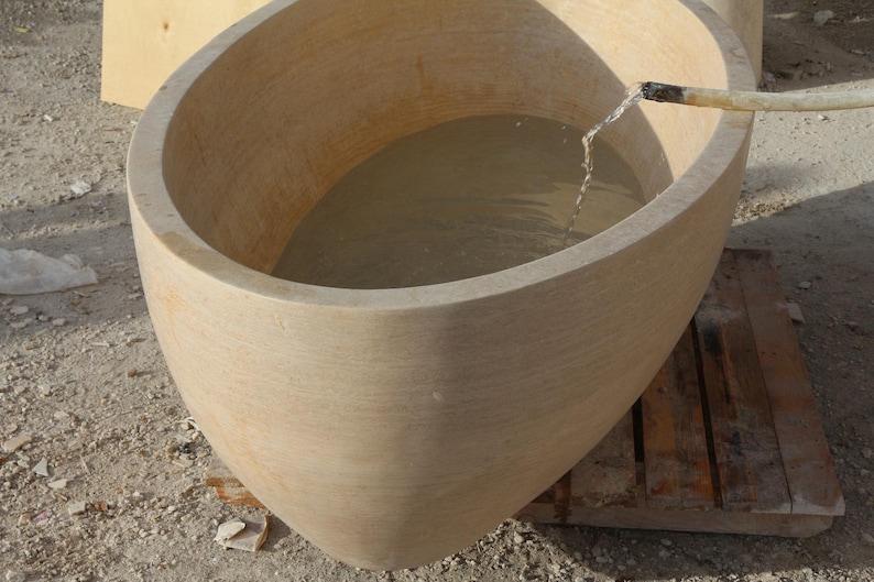 May include: A large, beige stone bathtub with an oval shape. Water flows into the tub from a hose. The tub sits on a wooden pallet, and the background is a construction site.