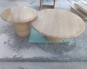 May include: Two round, light brown, stone coffee tables. The larger table is on a light blue mat. The smaller table is slightly raised on a pedestal.