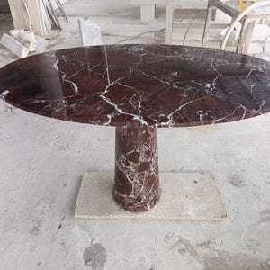 Può includere: Tavolo ovale in marmo con un motivo venato rosso scuro e bianco. Il tavolo ha un'unica base a piedistallo cilindrica e poggia su una base rettangolare. Le dimensioni del tavolo sono di circa 152 cm di lunghezza e 102 cm di larghezza.