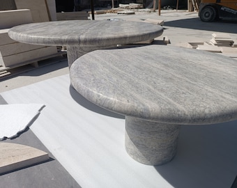 May include: Two gray stone tables with round tops and cylindrical bases. The tables have a smooth, polished surface with natural veining. The tables are made of a light gray stone and are set on a white surface.